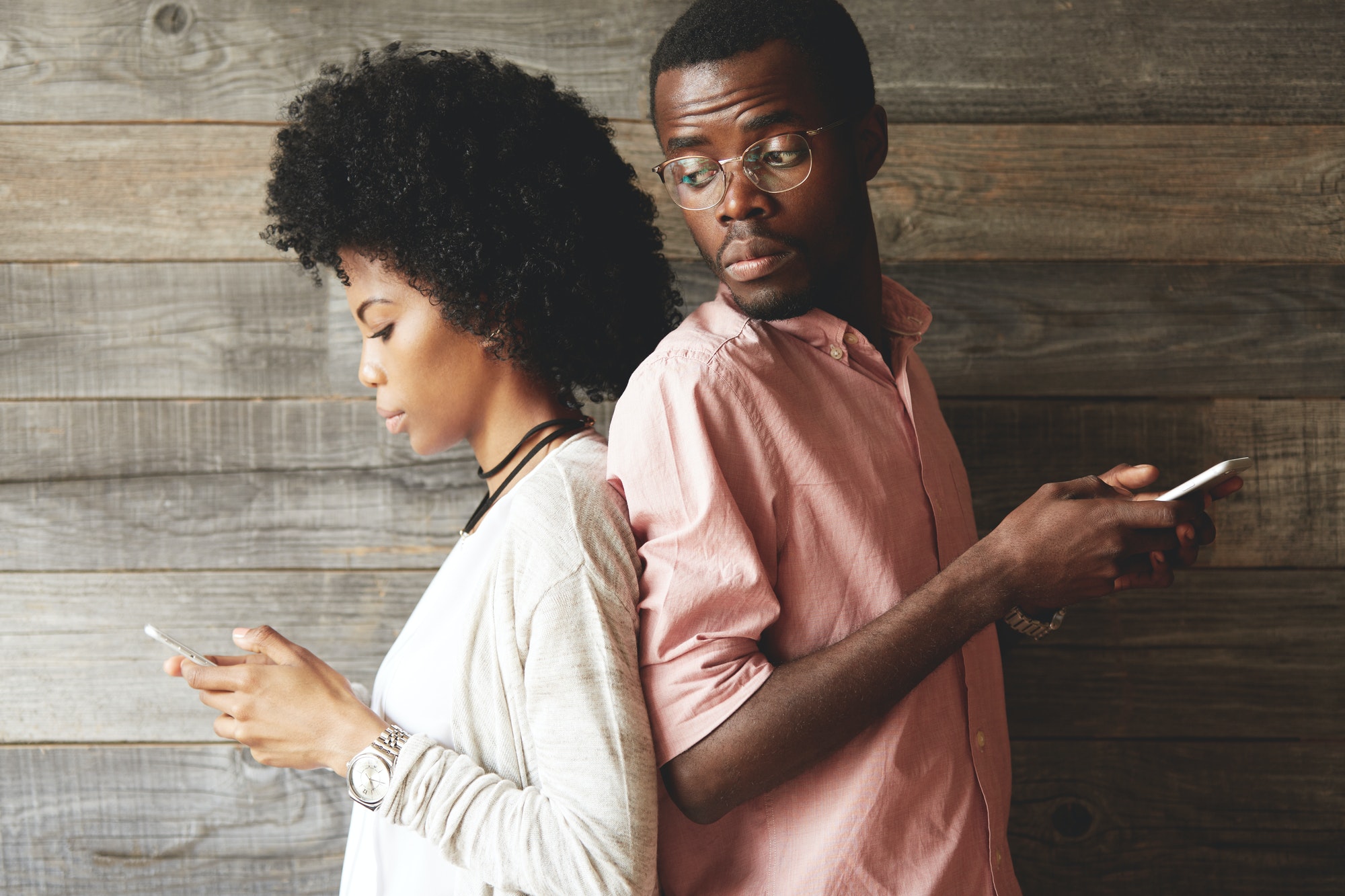 Young African couple standing back-to-back, holding mobile phones. Jealous possessive black man look